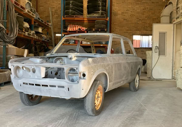 A dusty, partially disassembled 128 car inside a workshop with shelves and tools in the background.