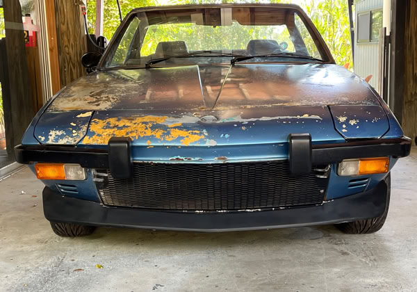 A weathered blue Fiat X1/9 convertible car with a rusted hood and visible paint damage, parked in a garage.