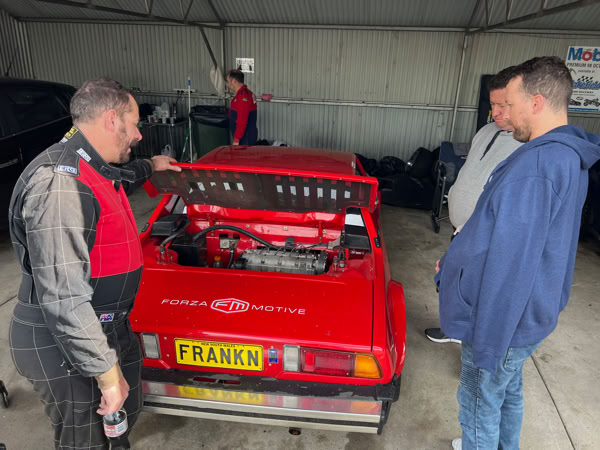 Club members inspecting the custom 16-valve engine bay of a modified Fiat X1/9 track car in the pits at One Raceway.