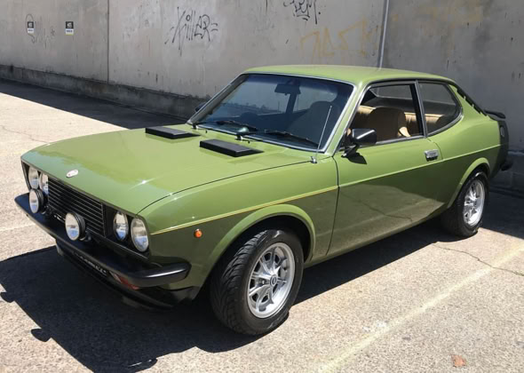 A green 128 car parked on a concrete surface, with a two-door coupe design and round headlights.
