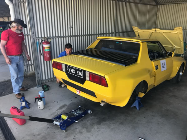 Fiat X1/9 with a Dallara wide-body kit jacked up in the pits for a brake pad inspection during a track day.